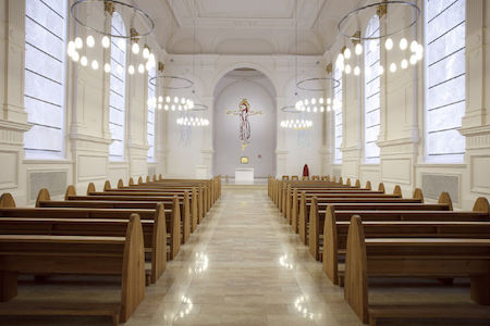 Heller, leerer Kirchenraum mit Holzbänken und einem Kruzifix am Altar.