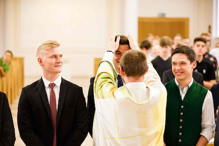 Ein Priester segnet einen lächelnden jungen Mann in einer Reihe mit anderen in Abendgarderobe.