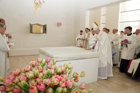 Priester in weißen Gewändern versammeln sich um ein Marmorgrab in einem hellen, weißen Raum.